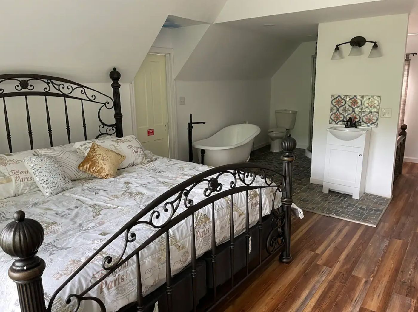 Bedroom under a sloped ceiling with a scrolled iron bedstead and a free-standing clawfoot bathtub.