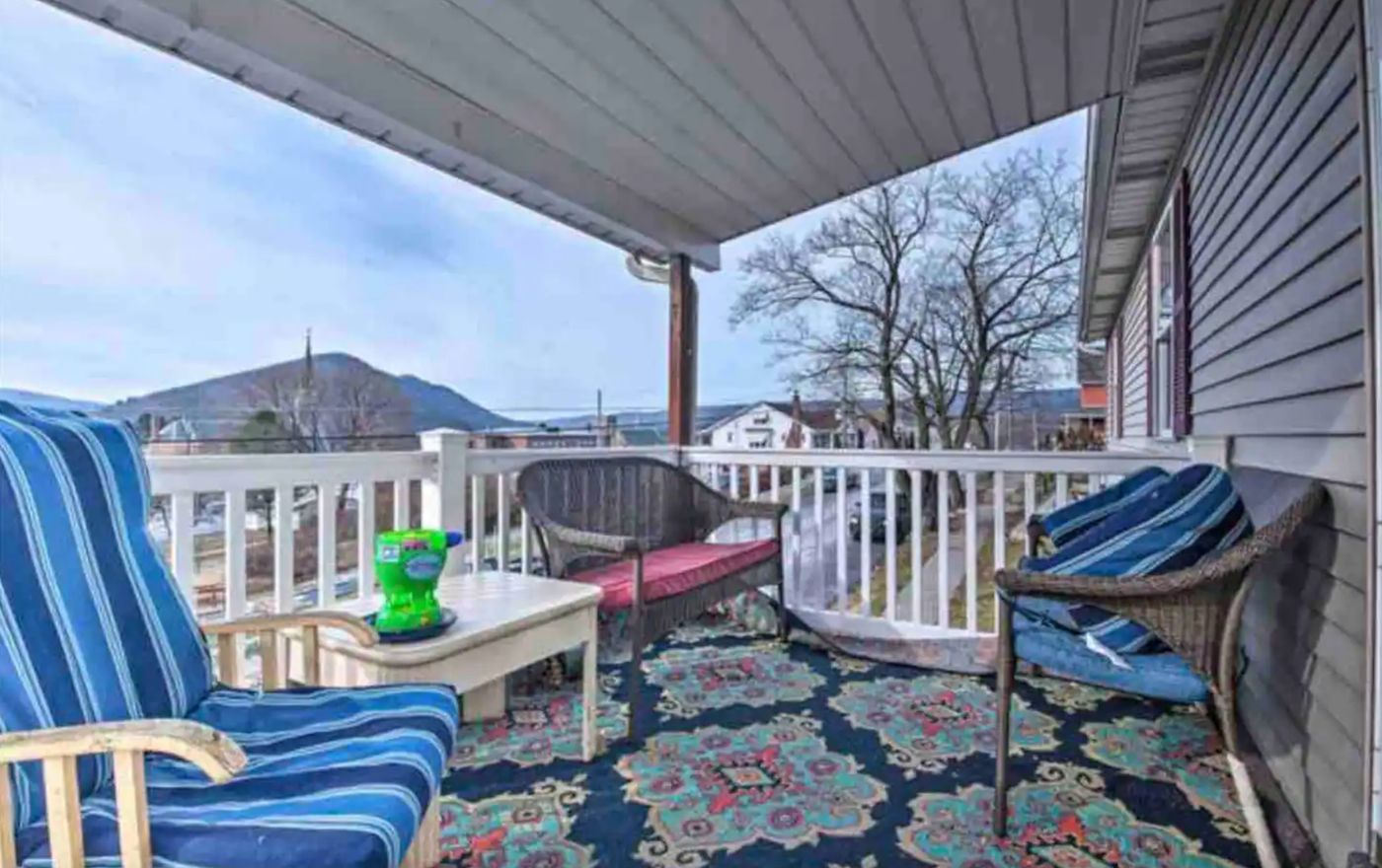 Covered front porch with striped lounge chairs and wicker seating, looking over rooftops to a wooded mountain.